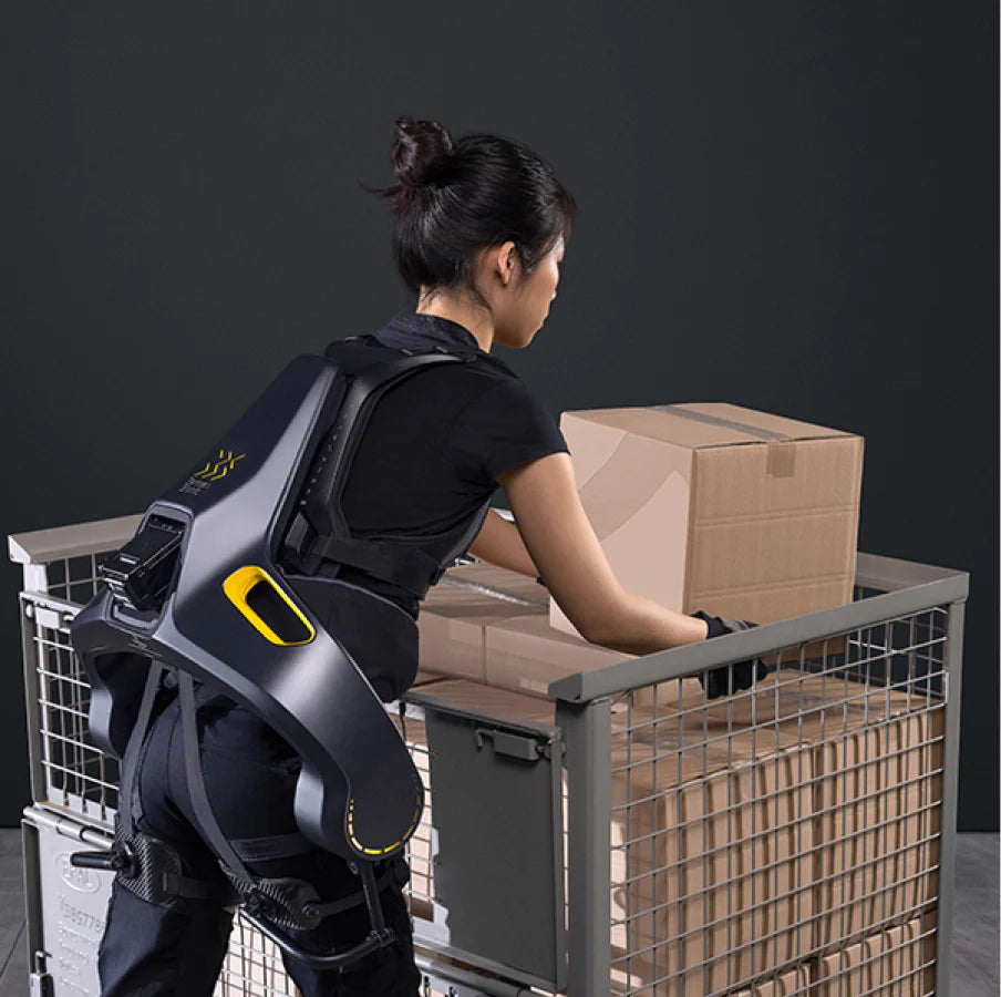 A woman wearing the German Bionic Apogee exoskeleton while lifting a cardboard box from a metal cart, demonstrating the exoskeleton’s ergonomic support in an industrial setting. – Now available at MTN Shop EU.