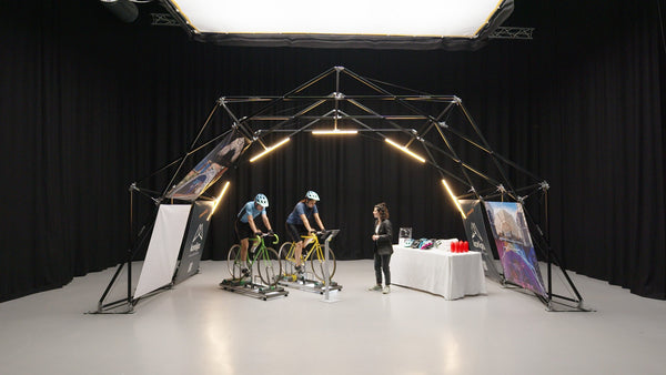 Indoor setup of Konligo event structure featuring two cyclists on stationary bikes and a display table with helmets and water bottles. Now available on MTN Shop US.
